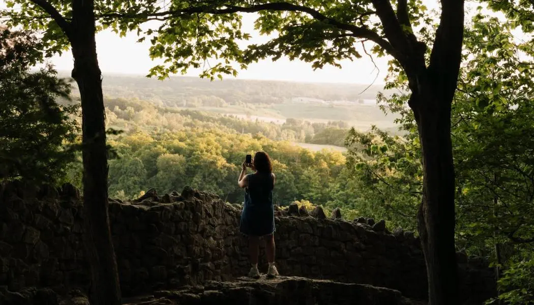 Una mujer se detiene en un mirador de piedra para tomar una fotografía del paisaje boscoso que se extiende a sus pies.