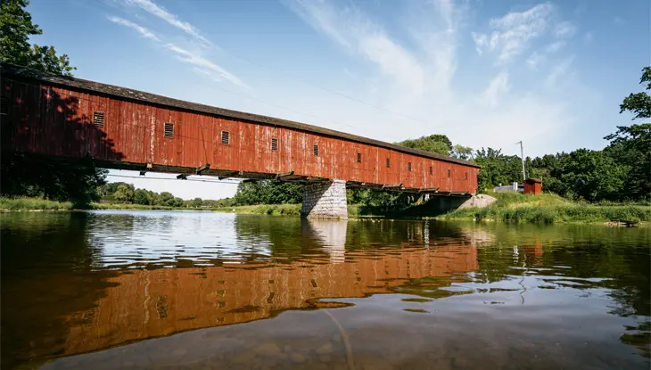 A red covered bridge crosses a tranquil river surrounded by green trees under a bright sky in West Montrose, Ontario.