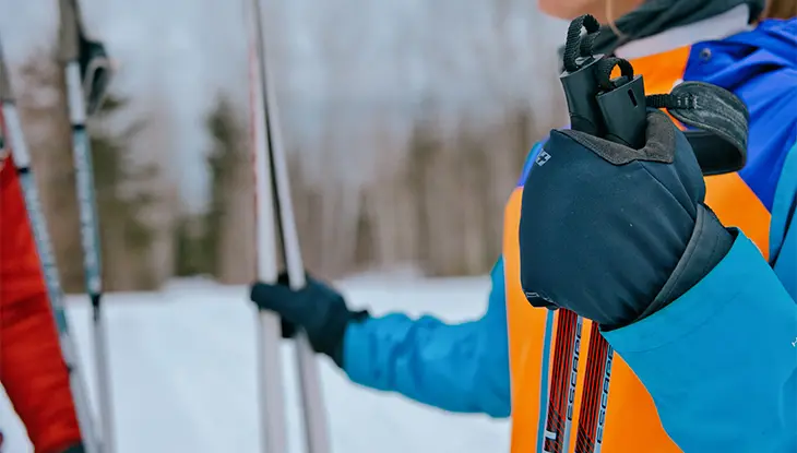 Vue rapprochée d’une personne portant des gants et un manteau orange, qui tient des bâtons de ski de fond dans la neige, près d’une forêt.