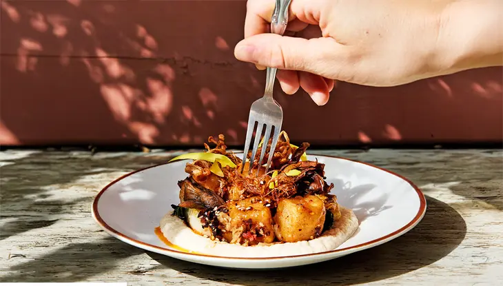 A hand holding a fork over a plated dish with crispy vegetables, golden cubes, creamy sauce and vibrant garnishes.