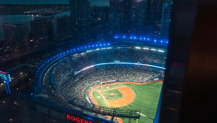 Vue aérienne en soirée d’un stade de baseball bien éclairé et bondé de spectateurs. On aperçoit la ligne d’horizon de la ville et le quartier riverain en arrière-plan.