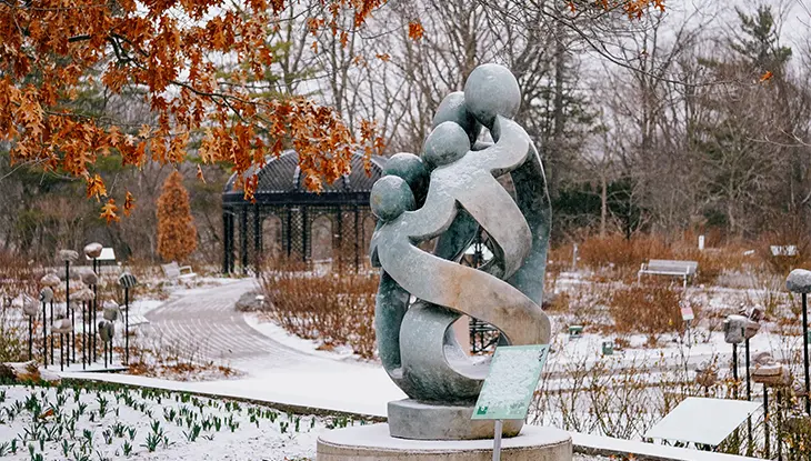 A metal statue located in the middle of the snow-covered garden depicts a small child embracing two adult figures.