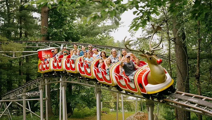 Personnes à bord d’un manège coloré dont le wagon avant est en forme de renne, dans un parc d’attractions boisé.