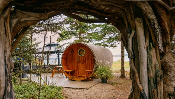 Vista a través de un arco de raíces de árboles que conduce a una pequeña sauna redonda de madera.