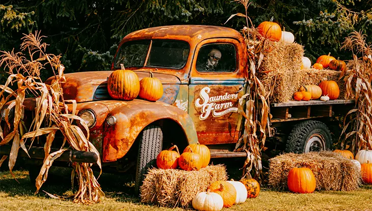Rusted vintage pickup truck decorated with pumpkins, hay bales and dried corn stalks for a fall harvest display.