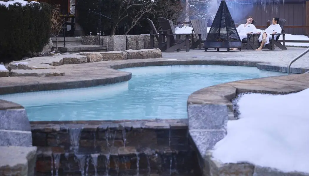 Zwei Personen in weißen Frotteemänteln wärmen sich im Winter an einem Feuer im Freien in einem nordischen Spa.