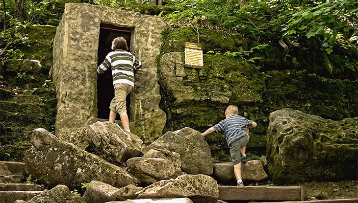 Deux enfants montant un escalier de pierre et s’approchant d’une entrée aménagée à flanc de montagne.