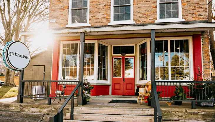 Historic village shopfront with a red door, large display windows and signage along a sidewalk.