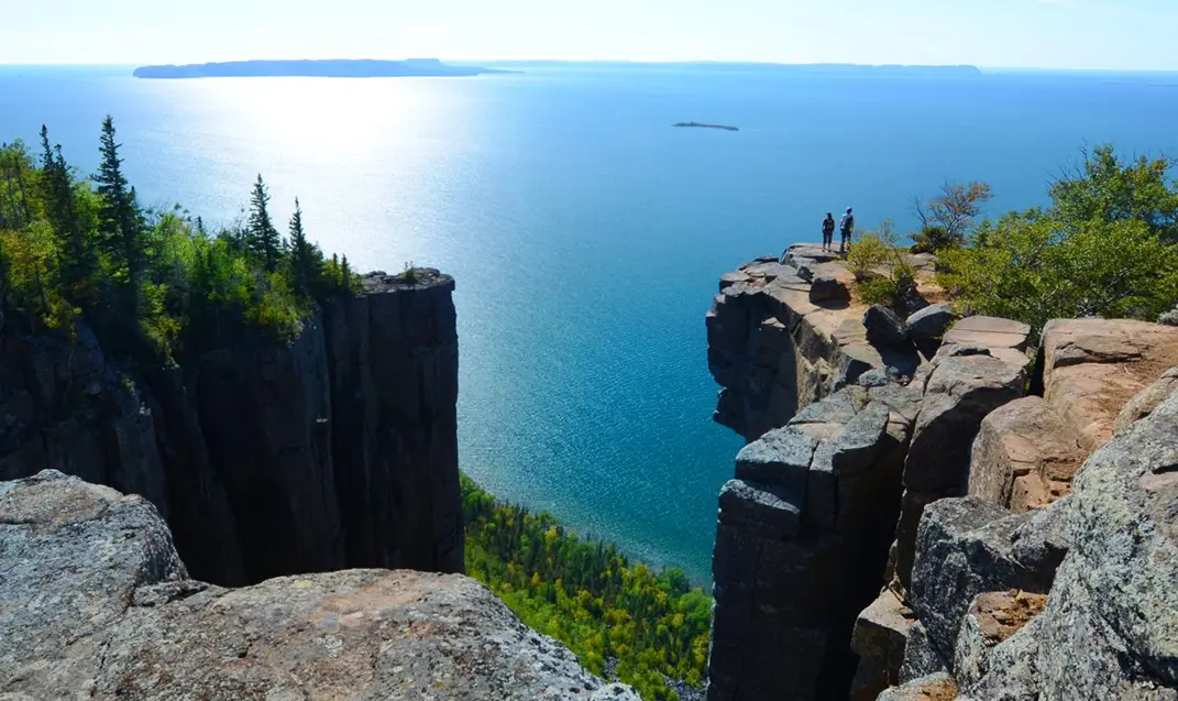Zwei Wanderer stehen am Rand einer Klippe auf dem Gipfel des Sleeping Giant und zeichnen sich als Silhouetten vor dem weiten Blau des Lake Superior ab.
