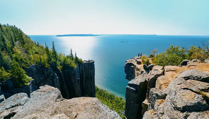 Two hikers on the edge of a cliff at the top of Sleeping Giant stand silhouetted against the vast blue of Lake Superior.