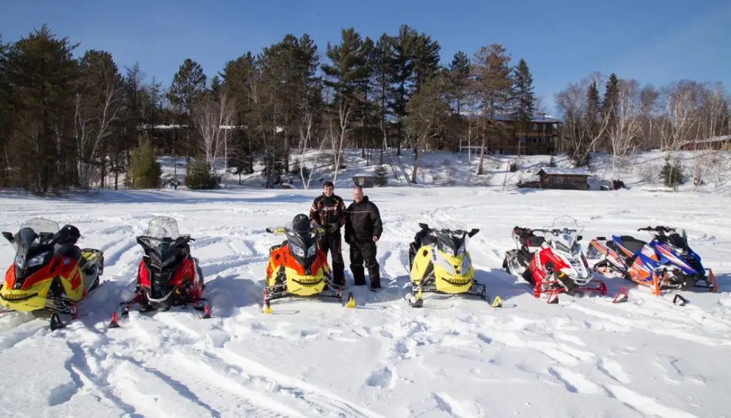 Riders with six snowmobiles parked on a frozen lakefront below cottages.