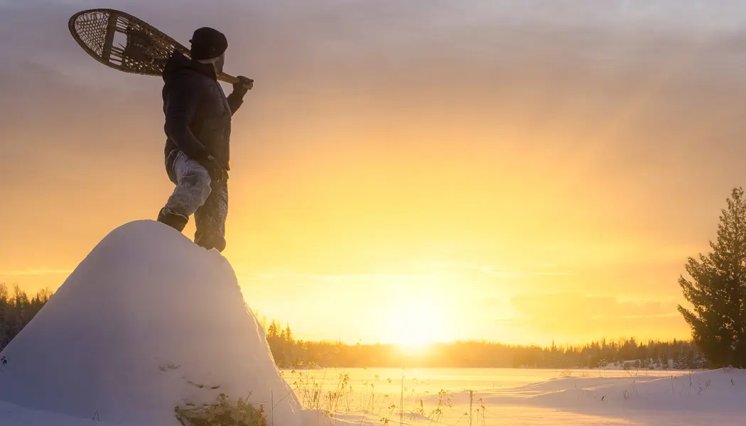 Ein Mann mit einem Paar Schneeschuhen steht im Schnee und beobachtet den Sonnenuntergang über einer gefrorenen Landschaft im Quetico Park.