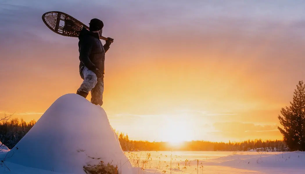 A man holding a pair of snowshoes stands in the snow watching the sun set over a frozen landscape in Quetico Park.