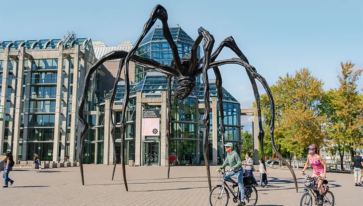 Large outdoor spider sculpture in an urban plaza with people walking and cycling nearby.