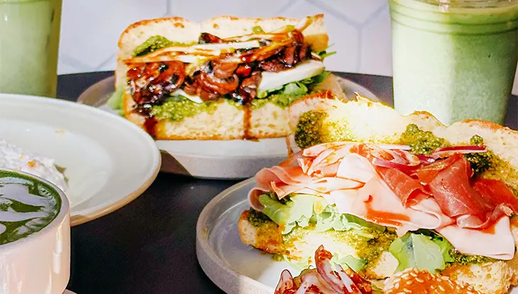 Sandwiches with cured meats and greens served with matcha drinks on a table.