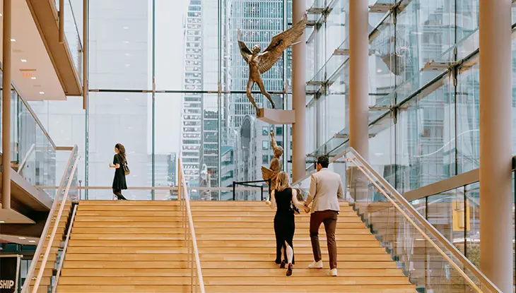 People walking up a wide staircase inside a modern glass atrium with tall windows and city views.