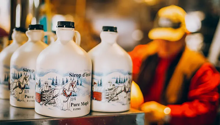 Maple syrup bottles lined up on a counter, with a worker blurred in the background inside a sugar shack.