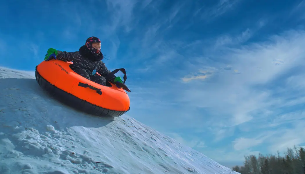 A child bundled up for the winter weather rides a bright orange tub down a snowy hill at a winter festival.