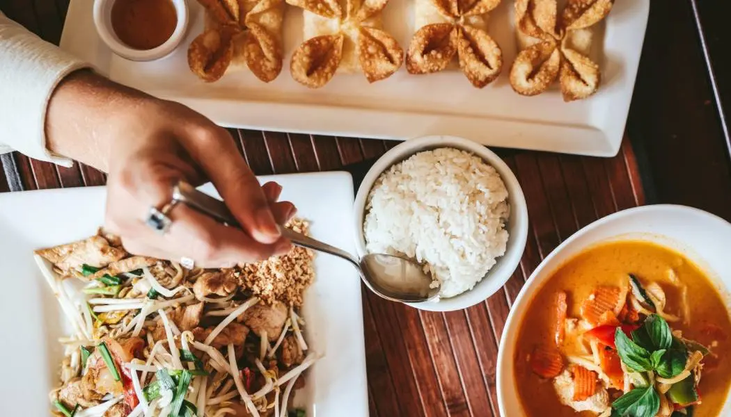 Une personne se servant du riz à la louche dans un plat entouré d'un bol de curry, de nouilles et de fritures.