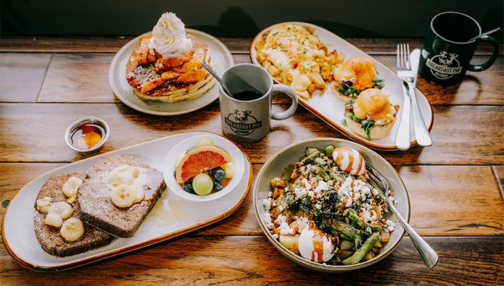Ein reichhaltiges Brunchbuffet mit Pfannkuchen, Eggs Benedict, French Toast mit Bananen und Kaffee auf einem Holztisch.
