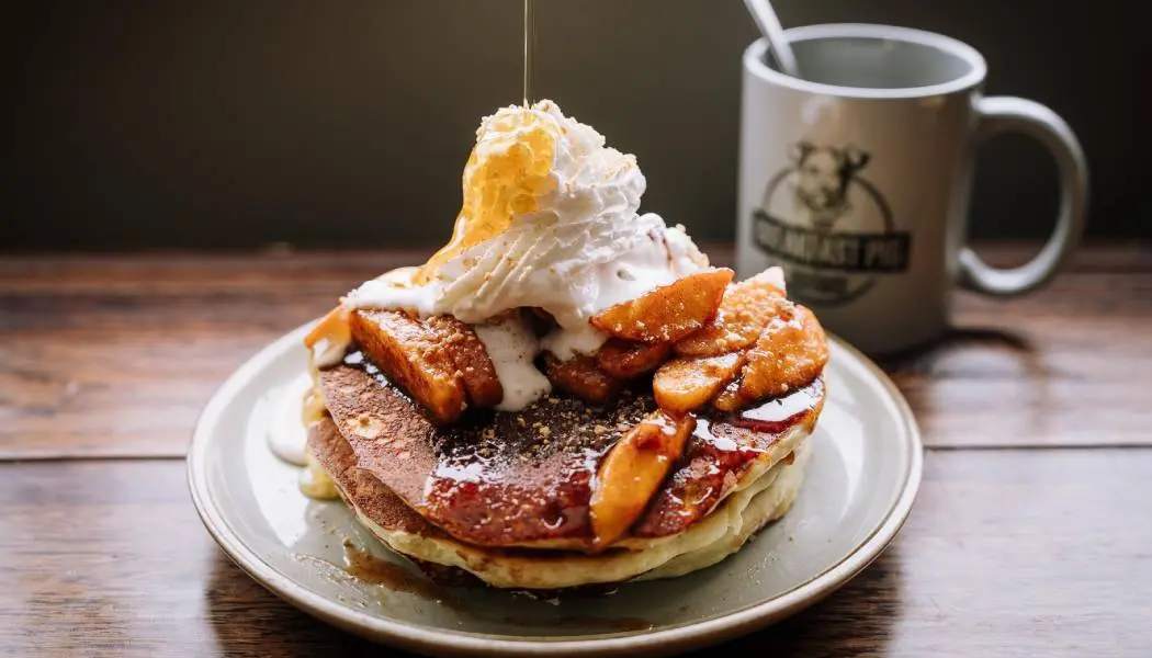 Du sirop d’érable est versé sur une assiette de crêpes garnies de fruits et de crème fouettée.
