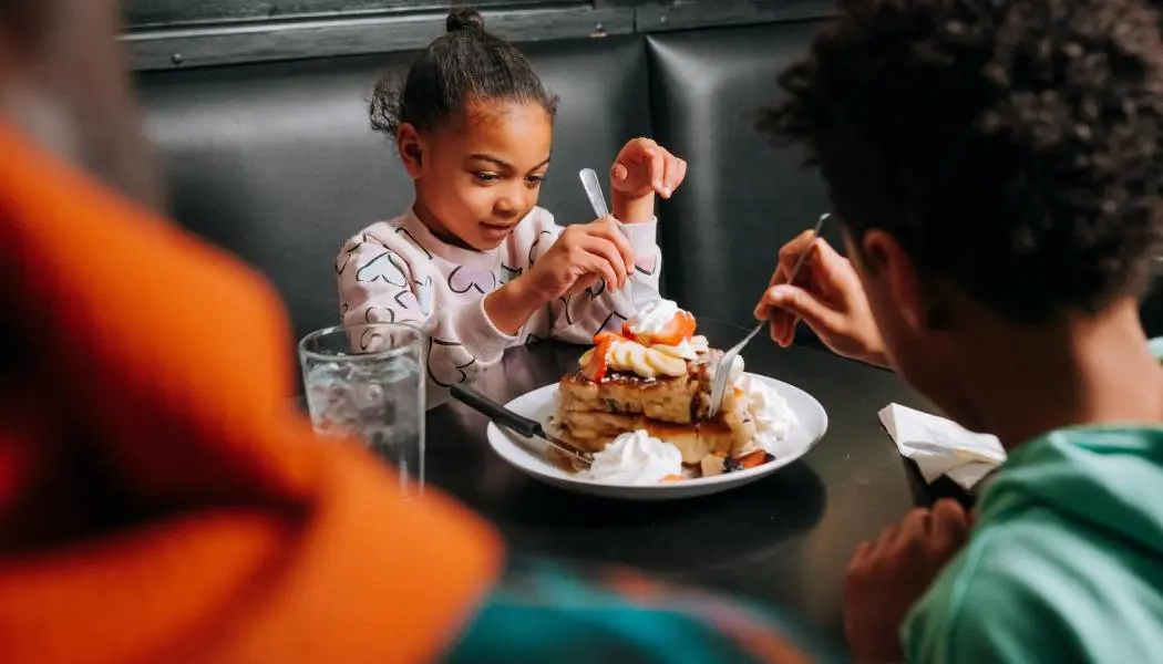 Deux jeunes enfants partagent une assiette de crêpes empilées, garnies de fruits et de crème fouettée.