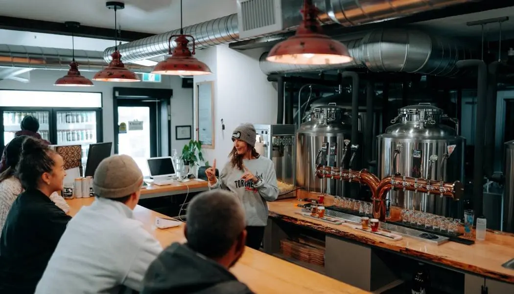A bartender takes the drink orders from a group of customers sitting at the wooden bar in a craft brewery taproom.
