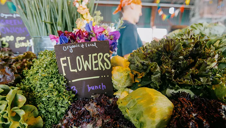 Un puesto en un mercado de agricultores que exhibe verduras de hoja verde y flores, con un cartel que dice "flores orgánicas y locales".