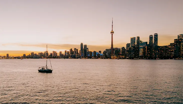 A single sailboat cruises the Toronto harbour at dusk with the CN Tower in the background.