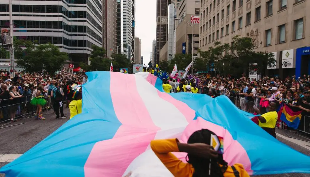 Groupe de personnes transportant un grand drapeau trans bleu, rose et blanc au centre de la rue Yonge, durant un défilé de la Fierté à Toronto.