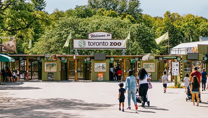 Visiteurs marchant vers l’entrée du zoo de Toronto, avec l’affiche de bienvenue au-dessus du portail et des arbres en arrière-plan.