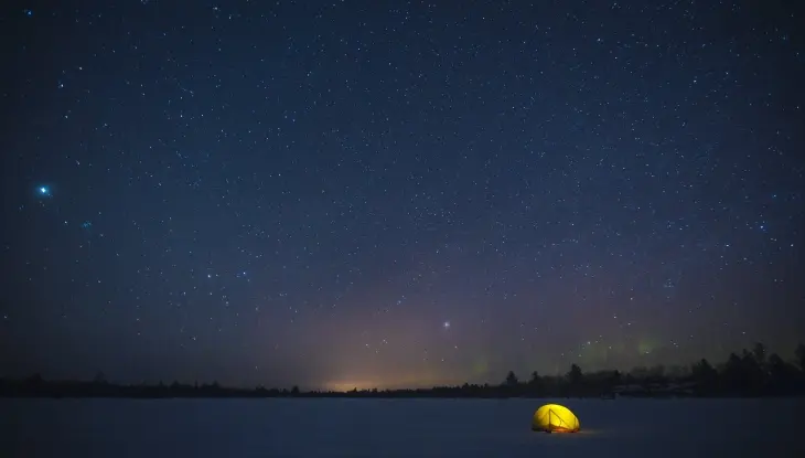 Ein nachts im Schnee beleuchtetes Zelt, im Hintergrund Bäume und Sterne am Himmel