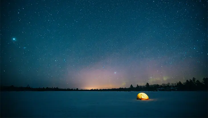 Tent lit up at night in the snow with trees in the background and stars in the sky.