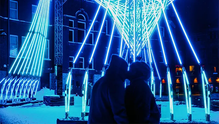 Couple standing in silhouette beneath a bright geometric light installation at night.