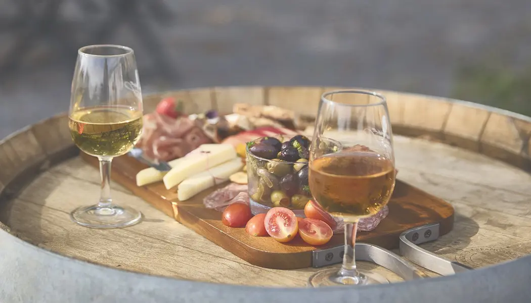 Two glasses of wine and a charcuterie of cheese, meats and olives sit on the wine barrel table.