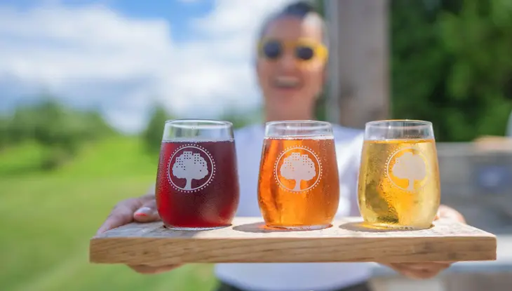 Una mujer sostiene una tabla de madera con tres sidras recién servidas para probar en West Avenue Cidery en Hamilton.