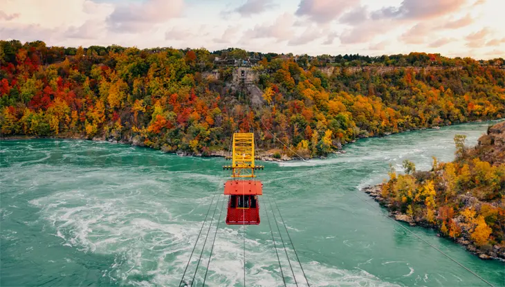 A red cable car travels across the Niagara River with stunning fall colour foliage in the background.