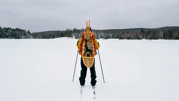 Eine Person fährt auf Skiern über einen zugefrorenen, schneebedeckten See und trägt dabei hölzerne Schneeschuhe auf dem Rücken; in der Ferne sind ein Wald und ein bedeckter Himmel zu sehen.