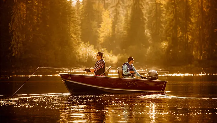 Two people fishing from a small boat on a calm lake surrounded by forest at golden hour.