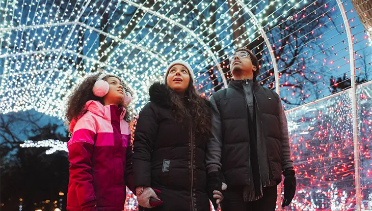 Two adults and a child look up in awe as they walk through a tunnel of white and blue twinkling holiday lights.